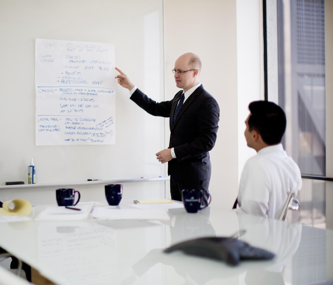 Two colleagues engaged in conversation in a conference room. One colleague is standing and points at words on a board and the other is seated..